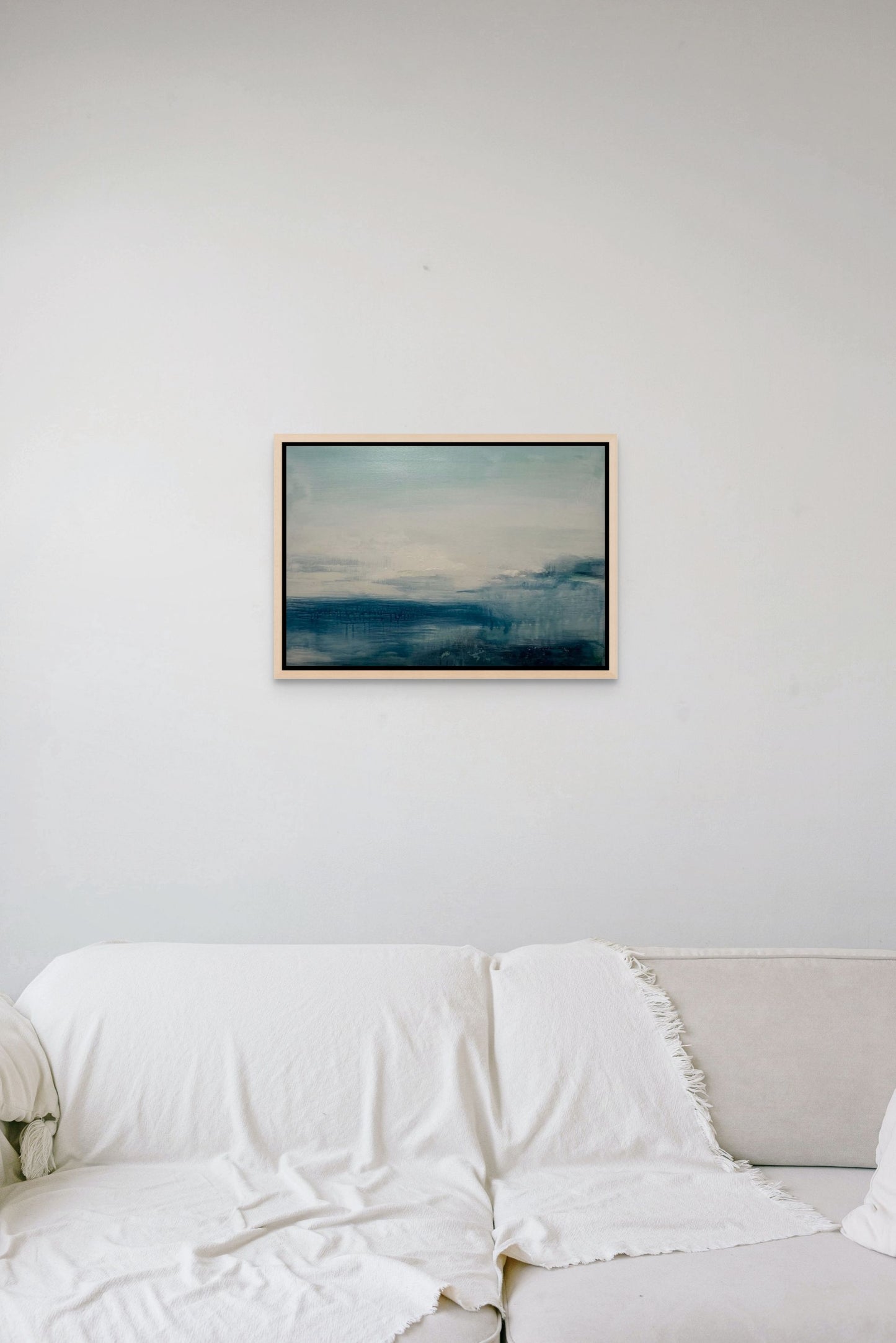 Framed abstract seascape artwork in grey and blue on a wall above a white sofa