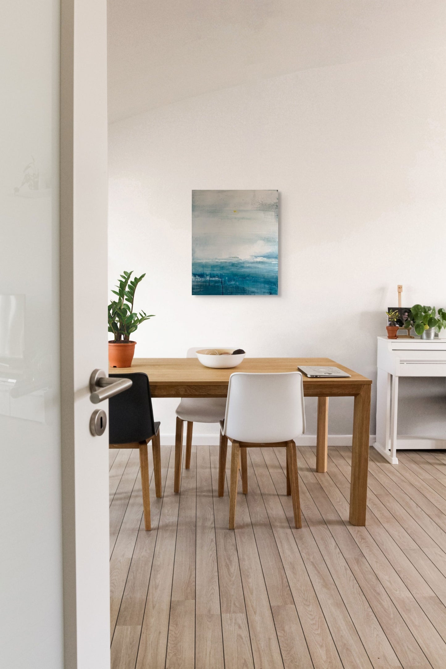 Dining area with wooden table and chairs, plants, and an abstract seascape painting on the wall.