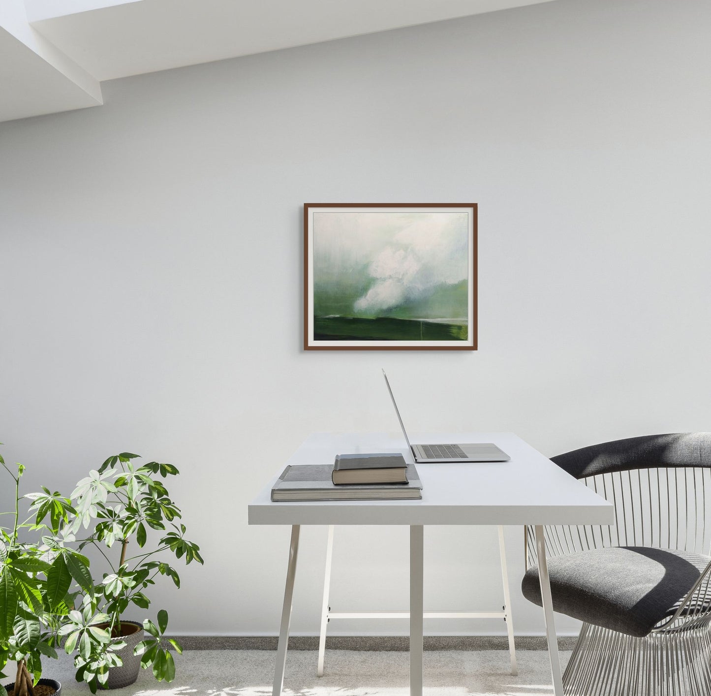 Framed artwork on a wall above a desk with a laptop and books in a room with a plant.