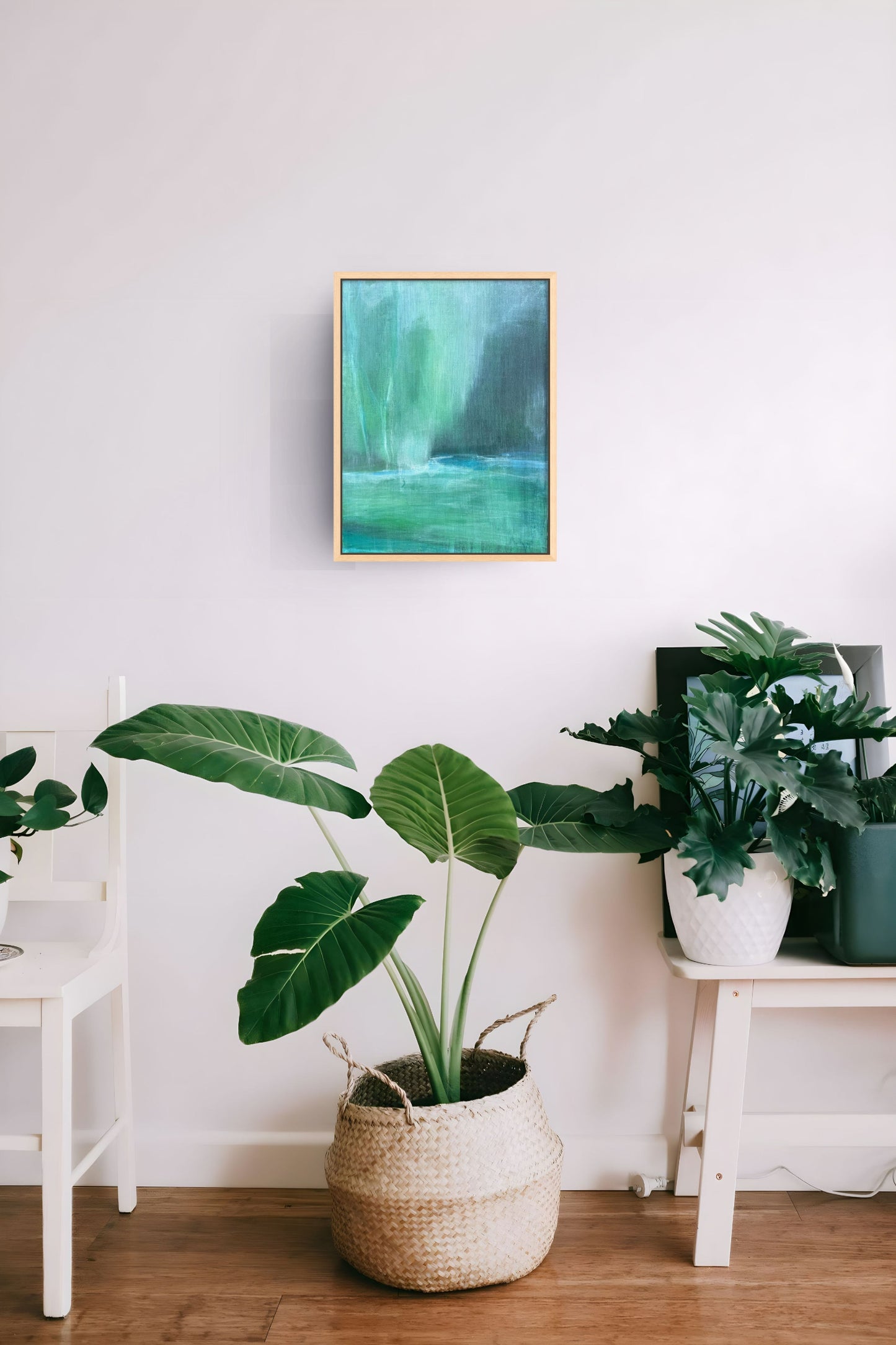 A framed abstract painting with green and teal tones hangs on a white wall above a potted green plant in a woven basket, with additional plants on white side tables in a bright interior setting.
