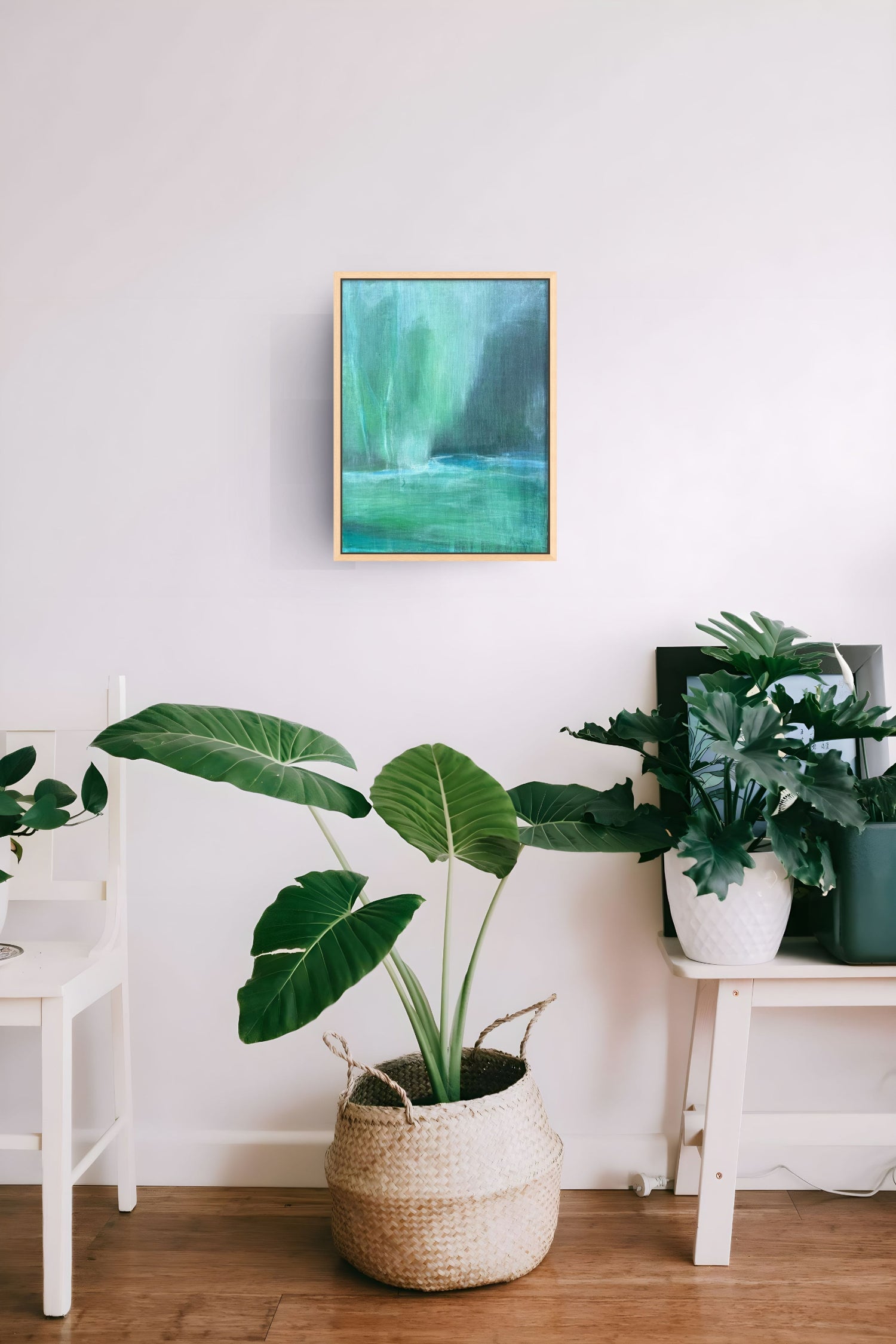 A framed abstract painting with green and teal tones hangs on a white wall above a potted green plant in a woven basket, with additional plants on white side tables in a bright interior setting.