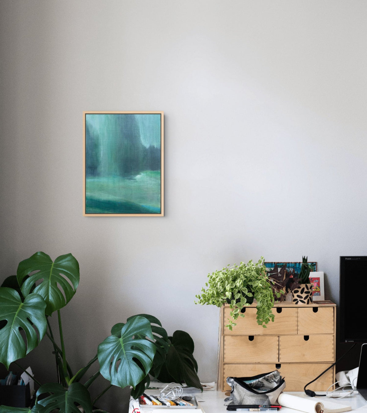 A framed abstract painting with teal, green, and blue tones hangs on a light gray wall in a styled room setting with a wooden dresser, plants, and desk accessories.