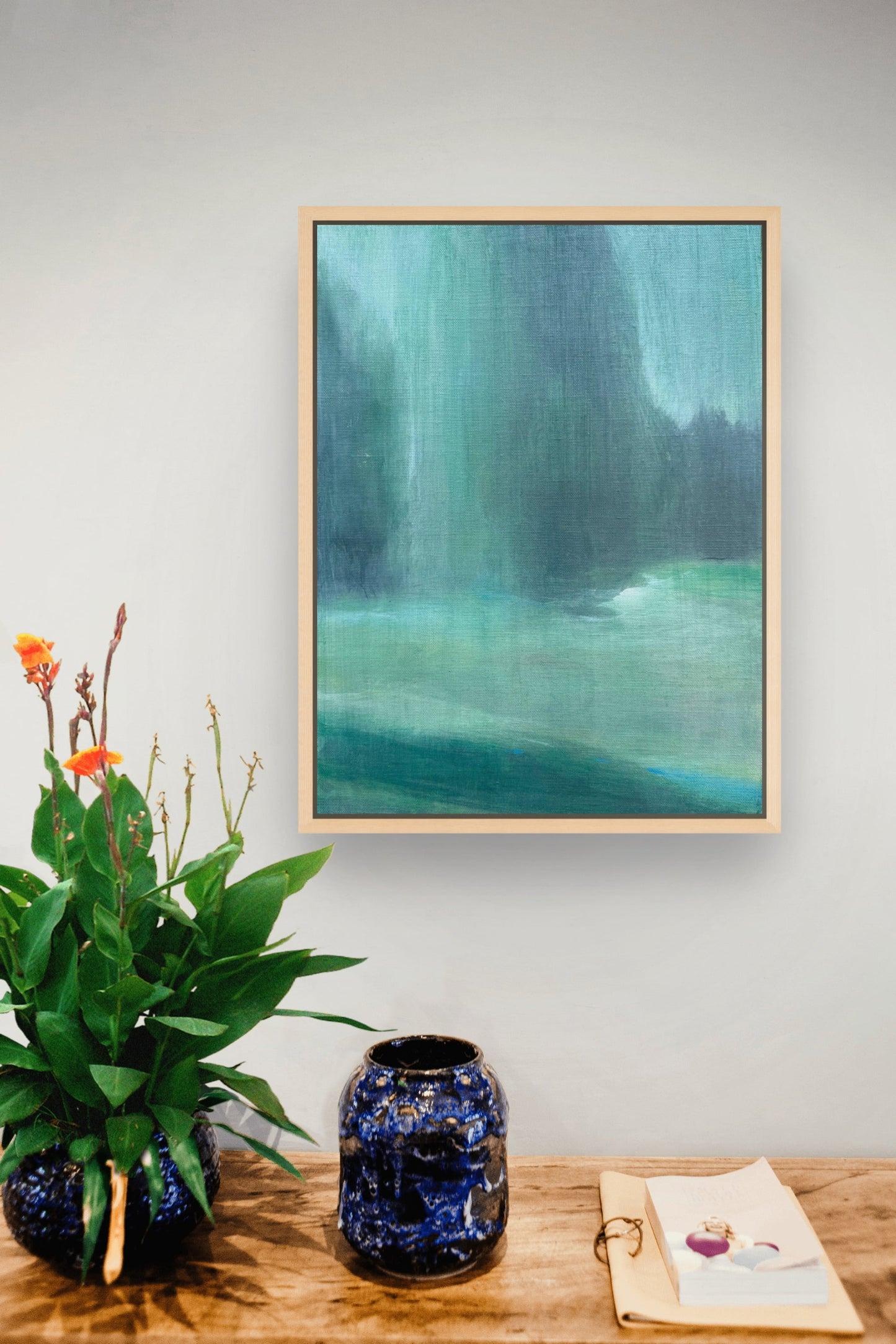 A framed abstract landscape painting featuring shades of teal, green, and blue, hanging on a light gray wall above a wooden table styled with a potted plant and a blue ceramic vase.