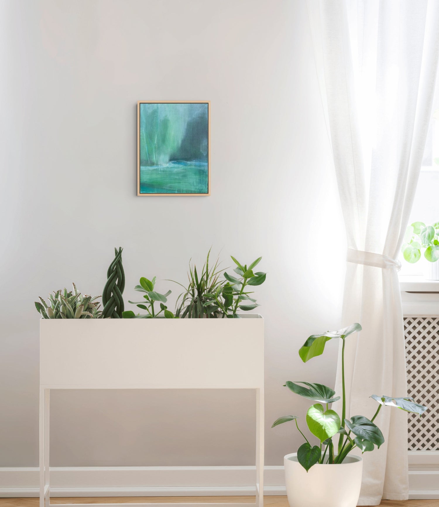 A small framed abstract landscape print with green and teal tones hangs on a light gray wall above a white planter box with several green plants. A potted plant sits near a window with sheer white curtains.
